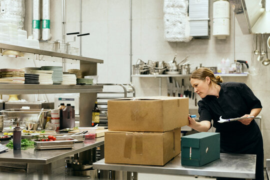 Female chef black uniform checking delivery boxes in commercial kitchen