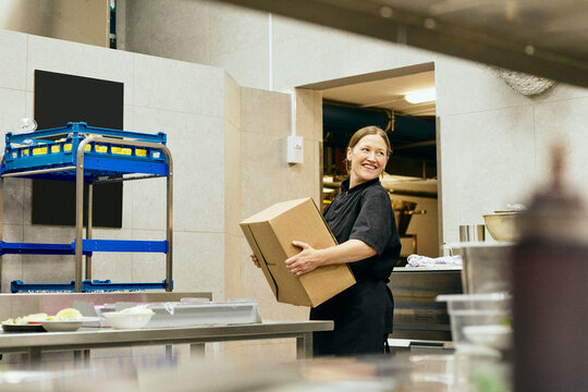 Smiling female chef carrying cardboard box through doorway
