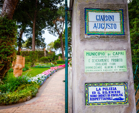 07 02 2026 - Capri, Italy. Entrance to Giardini Augusto, featuring a signboard display in foreground with a concrete and brick pathway adorned by vibrant flowers and trees, set against a lush green