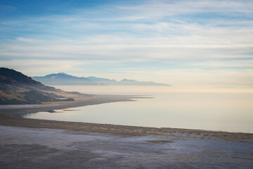Fototapeta premium Minimal travel background featuring a curved shoreline, perfectly still water, and layers of distant mountains covered in soft haze. High-key serene landscape at Antelope Island, Utah.