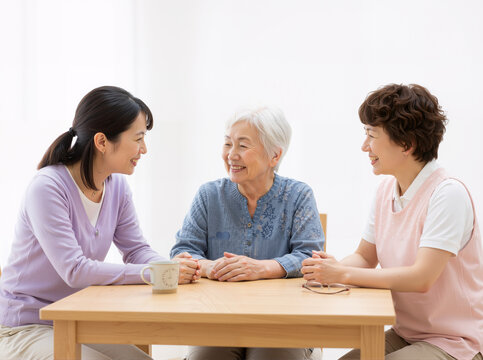 Three women share warm conversation: elderly woman centered, two younger women supporting her. Hands on shoulders show deep care and connection. Soft lighting, casual clothes, coffee cups create intim