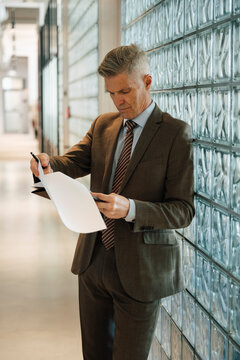 Senior businessman reading documents while leaning on glass brick wall at office