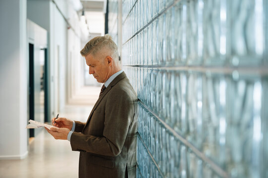 Side view of senior businessman analyzing reports while leaning on glass brick wall