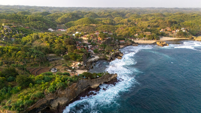 Aerial view of Baron Beach with its rugged cliffs, crashing waves, and lush green hills overlooking the coastline in Tanjungsari, Special Region of Yogyakarta, Indonesia.