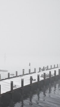 A dock stretches into fog covered water. The scene shows a deserted area with little visibility. Wood planks are visible, and the water reflects the gray surroundings.