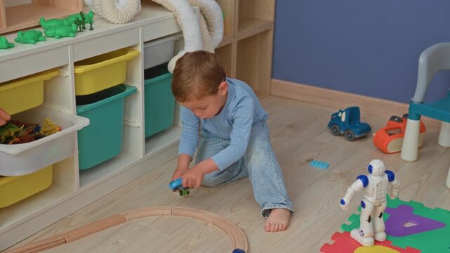 Boy kneeling pushing a toy car beside a blue white robot on foam puzzle mat near train track and storage shelf in building; playful curiosity.