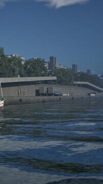 A boat floats on the calm river water near a concrete area. Buildings rise in the background against a clear blue sky. Sunlight reflects off the water surface.