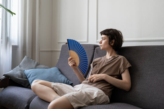 Weary tired woman struggling with sweltering weather using fan to cool down in airless room in summer. Heat exhaustion, physical fatigue, lack of ventilation, heatwave issues, urban living hardship