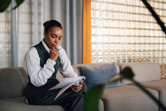Female entrepreneur with hand on chin examining documents while sitting on couch