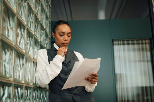 Thoughtful businesswoman reading documents while leaning on glass brick wall