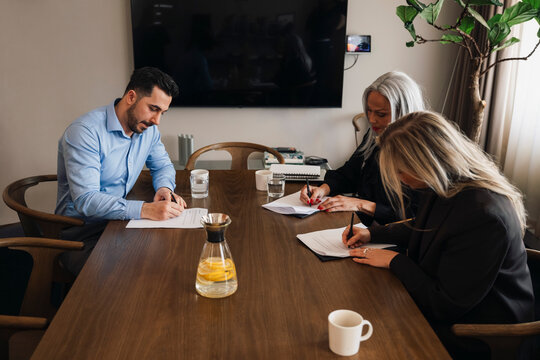 Male and female business professionals filling contract forms while sitting at wooden table in board room