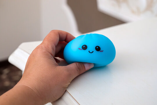 Hand holding a squishy blue dumpling toy with a cute smiling kawaii face and pink cheeks resting gently on a soft white wooden shelf indoors near a blurred wall