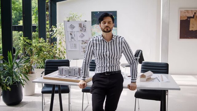 Bearded architect designer in striped shirt smiles leans on desk