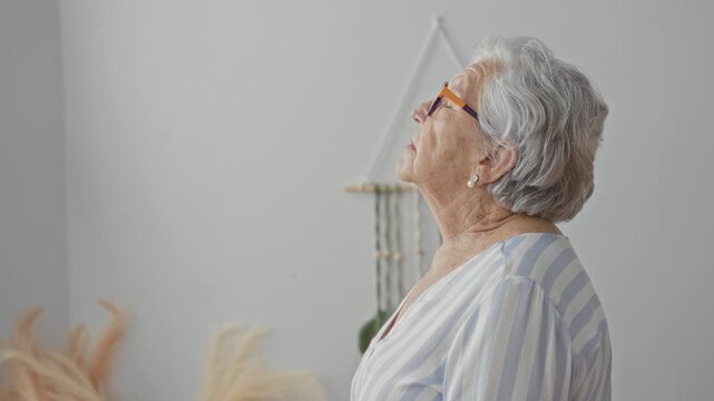 Woman wearing orange framed glasses tilts head back and gazes upward, pearl earring and bare neck visible, striped blouse by macrame wall hanger in a bedroom; serenity.