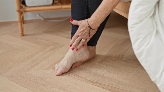 Woman in black leggings touches bare foot and ankle on light wooden parquet floor beside bed in building, with red nails, ring and bracelet; concern care.