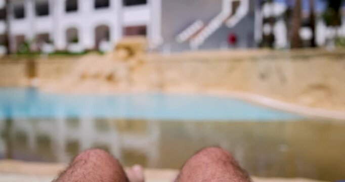 First person slow motion view relaxing beside a luxury swimming pool with feet bent up white sun lounger beside clear blue water and wooden decking at a tropical holiday resort during summer travel