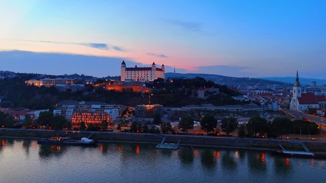 Bratislava Castle illuminated at dusk over the Danube River. Aerial view of the fortress on a hill with pink sunset sky and city lights.