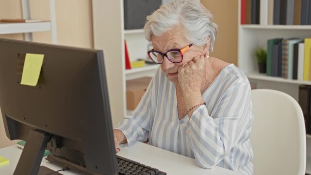 Woman at computer with hand on cheek looking at monitor, wearing eyeglasses and striped blouse, sticky note on screen in building; calm contemplation.