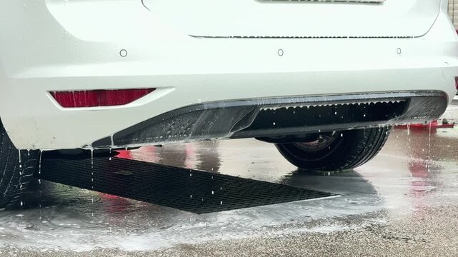Soapy water drips from rear bumper of vehicle over drainage grate at cleaning station. Car wash station, high-pressure water sprayer, manual vehicle detailing.