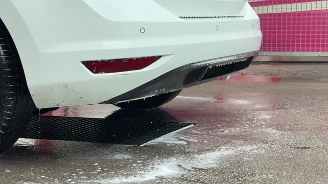 Soapy water drips from rear bumper of vehicle over drainage grate at cleaning station. Car wash station, high-pressure water sprayer, manual vehicle detailing.