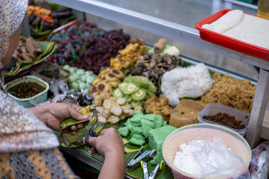 A high-angle close-up of a market vendor's hands preparing lenjongan, a traditional Javanese sweet snack assortment, on banana leaves at Pasar Gede, Solo.