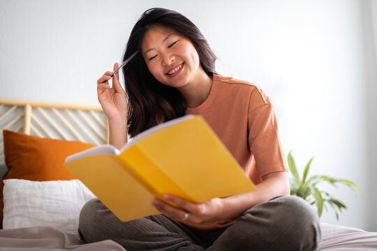 Asian woman writing journal smiling brainstorming ideas in bed