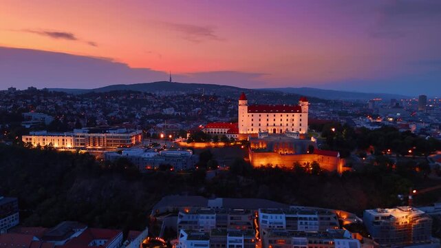Bratislava castle illuminated at vibrant sunset, Slovakia. Aerial view of stone fortress on hill with orange sky backdrop.