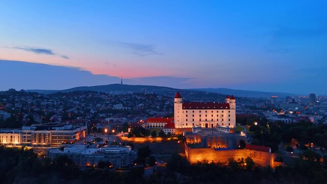 Bratislava castle at dusk with city lights, Slovakia. High angle shot of lit fortification building against twilight sky.