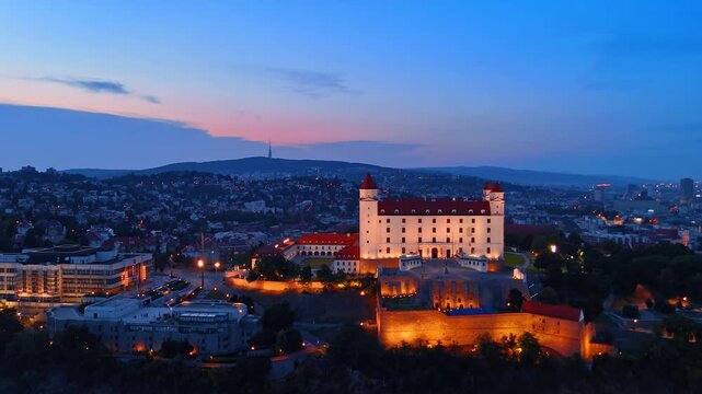 Bratislava castle overlooking Danube river at night, Slovakia. Aerial perspective of glowing palace building near illuminated bridges.