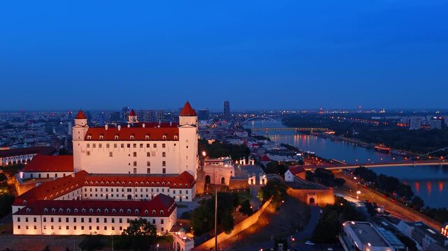 Bratislava castle walls glowing under night sky, Slovakia. Panoramic drone shot of bright white fortress walls at twilight.