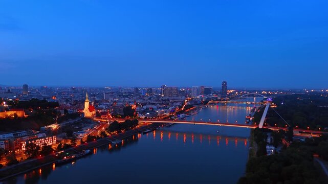 Aerial view of Bratislava skyline and Danube at twilight. Illuminated buildings and SNP Bridge reflected in water under deep blue evening sky.