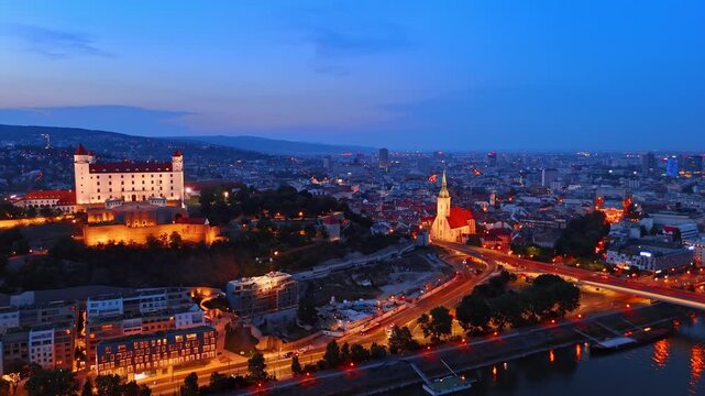 Aerial view of Bratislava Castle and historic town at night. Illuminated fortification on hill next to St Martins Cathedral and bright city street lights.