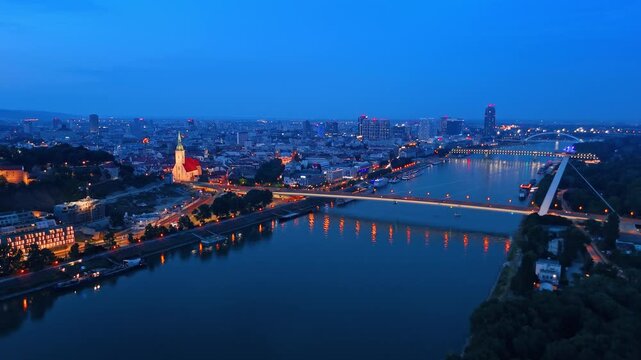 Drone panorama of Bratislava cityscape along Danube river during twilight. Night illumination on city buildings and bridges with dark blue sky background.