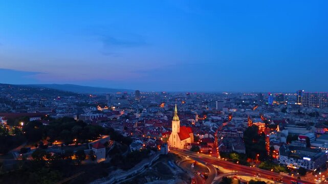 Evening drone cityscape of Bratislava old town and cathedral spire. Elevated overview of historical European streets with glowing traffic lights during dusk.