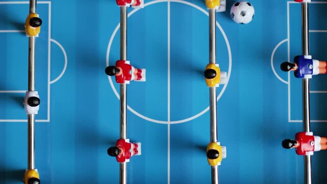 4K Slow motion video of a table football foosball match from above. Top view flat lay shot of miniature red and yellow plastic soccer players on metal rods moving a small ball on a blue field.