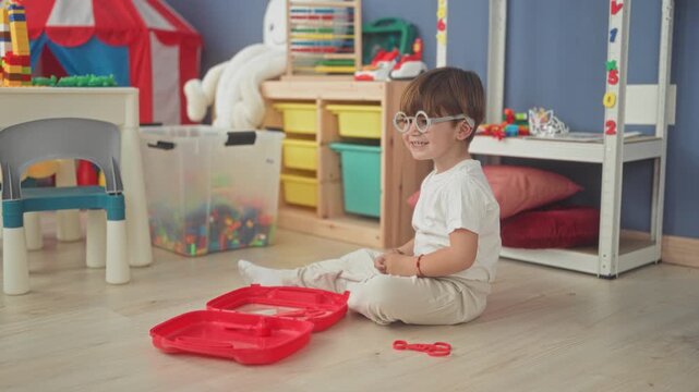 Boy wearing round glasses sits crosslegged on floor playing with a red toy case and plastic tongs in a playroom inside a building; playful joy.