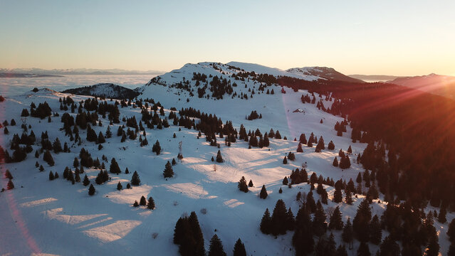 Aerial view of the snowy slopes of Station Monts Jura - site de La Faucille at sunset, with pine trees and golden light in Mijoux, Auvergne-Rhone-Alpes, France.