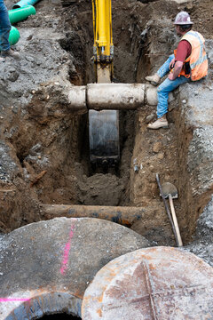 Excavator digging a deep narrow ditch in the dirt to work on underground utilities, construction worker in trench guiding machine operator around old concrete pipe
