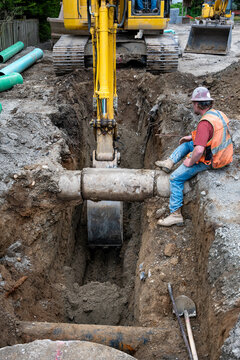 Excavator digging a deep narrow ditch in the dirt to work on underground utilities, construction worker in trench guiding machine operator around old concrete pipe

