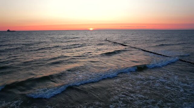 Cinematic drone shot moving forward over sea waves towards a wooden breakwater during a golden sunrise. A distant ship silhouette is visible on the pink horizon line in Chalupy, Hel Peninsula.