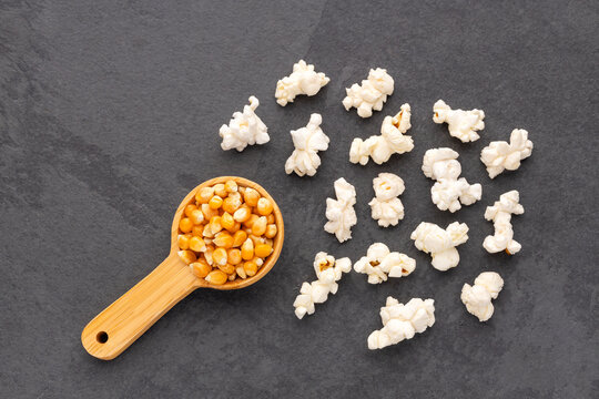 Wooden spoon with popcorn kernels next to popped popcorn on a dark stone background