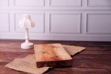 Soft glow warms wooden table's surface. Minimalist lamp stands beside rustic wood plank. Jute cloth adds texture beneath the wood. White wall paneling frames the tranquil scene © Nurma