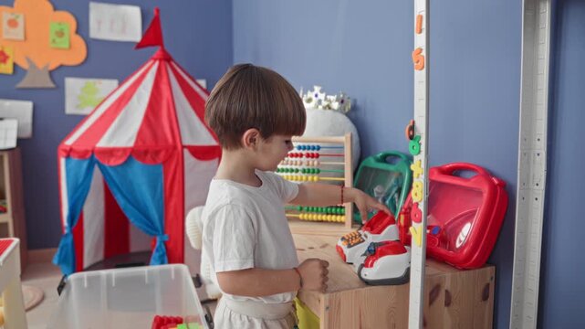 Boy presses toy register buttons and turns abacus beads beside a striped play tent and red toy case inside building; curiosity and learning.