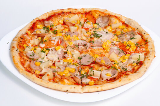 Whole baked pizza with sausages corn, tomatoes pineapple and herbs served on a white plate.