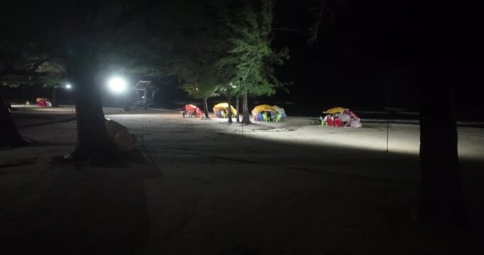 Very windy night shot: bright lights illuminate groups of people gathered under colorful parasols on a sandy beach, with waves crashing in the background.