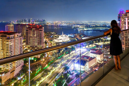 A young woman stands on a high rise terrace at night overlooking the illuminated Doha Pearl, downtown skyline, and the waters of the Persian Gulf in Doha Qatar.