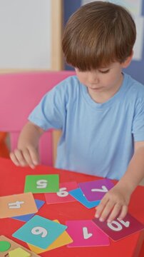 Boy placing numbered flashcards with both hands on a red table, arranging colorful counting cards and wooden shape puzzle pieces inside a preschool building; concentration learning.
