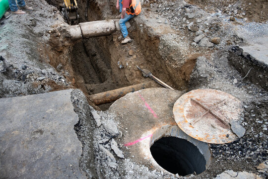 Underground utilities construction project, precast concrete stormwater system access vault in dirt trench under road, shovel and tamper bar, workman in trench guiding excavator around concrete pipe