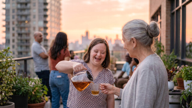 Adult daughter with Down syndrome pouring tea to mature mother during community sunset rooftop picnic