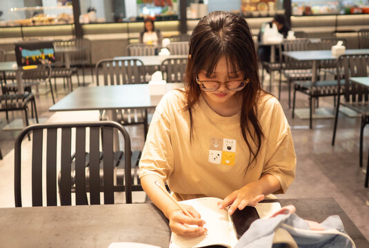 Young Asian female student wearing glasses focusing on writing in her notebook and using smartphone at a cafe. Gen Z lifestyle, education, and freelance remote working concept.